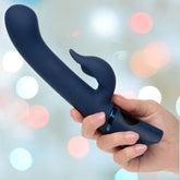 A hand holds the Orgasmatron Digital G-Teaser by CalExotics—a dark blue silicone vibrator with a curved tip and a smaller extension—set against a blurred backdrop of pastel lights.